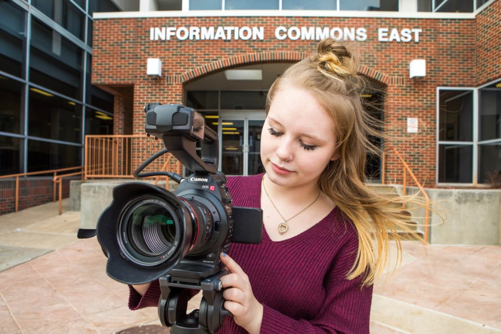 An electronic media production student taking a picture with a camera in front of the OTC Information Commons building